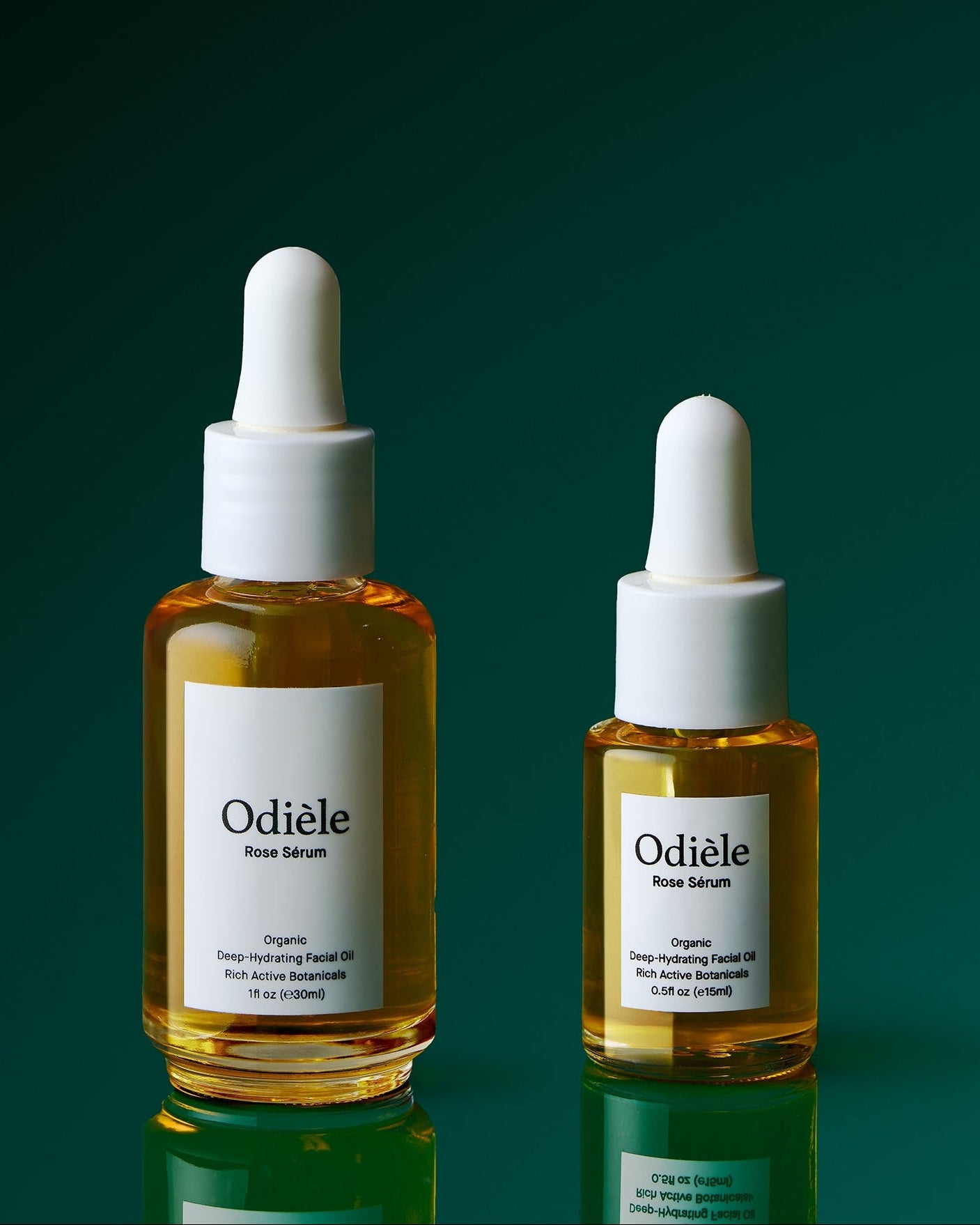 Two glass dropper bottles of Odiele Face Serum in a golden-yellow oil, displayed on a reflective dark green surface against a dark green gradient background.