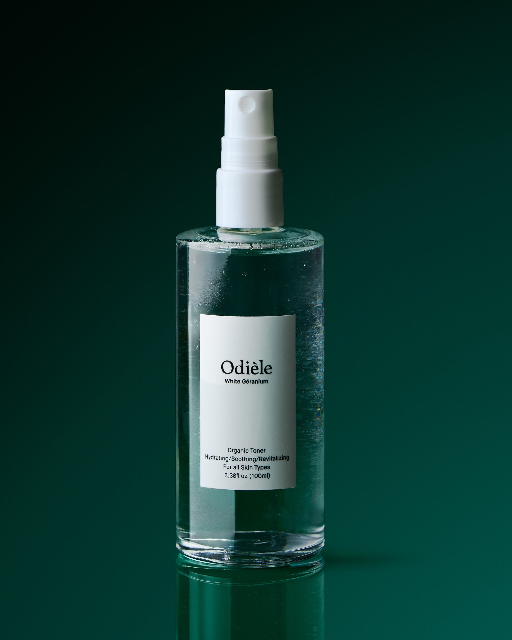 A clear glass spray bottle of Odiele Organic Tower hydrating moisturizing balmskin with a white pump and white label, set against a dark green gradient background.