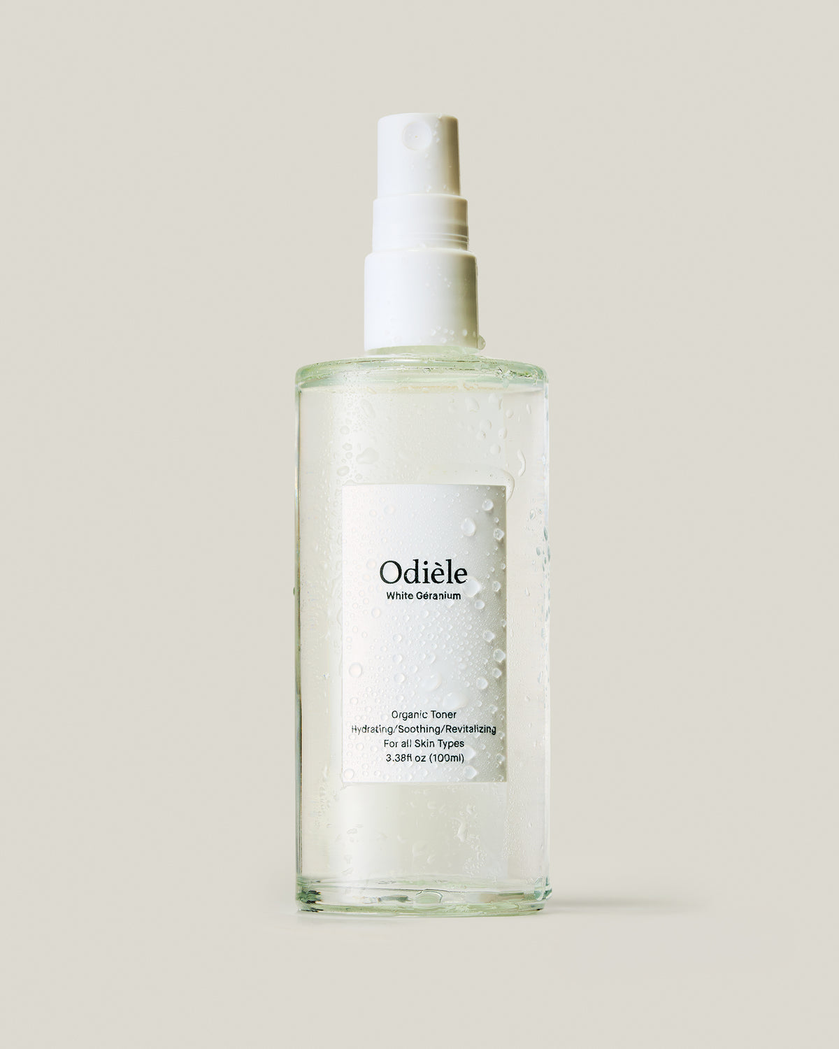 A clear glass spray bottle of Odiele Organic Toner with a white spray top and a white label, covered in water droplets, set against a solid light beige background.