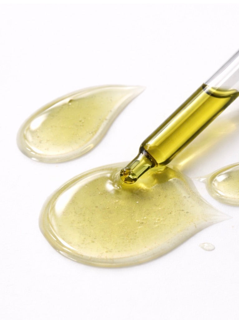 A clear glass dropper filled with pale yellow oil, dispensing into glossy pools of the same translucent yellow liquid on a white background.