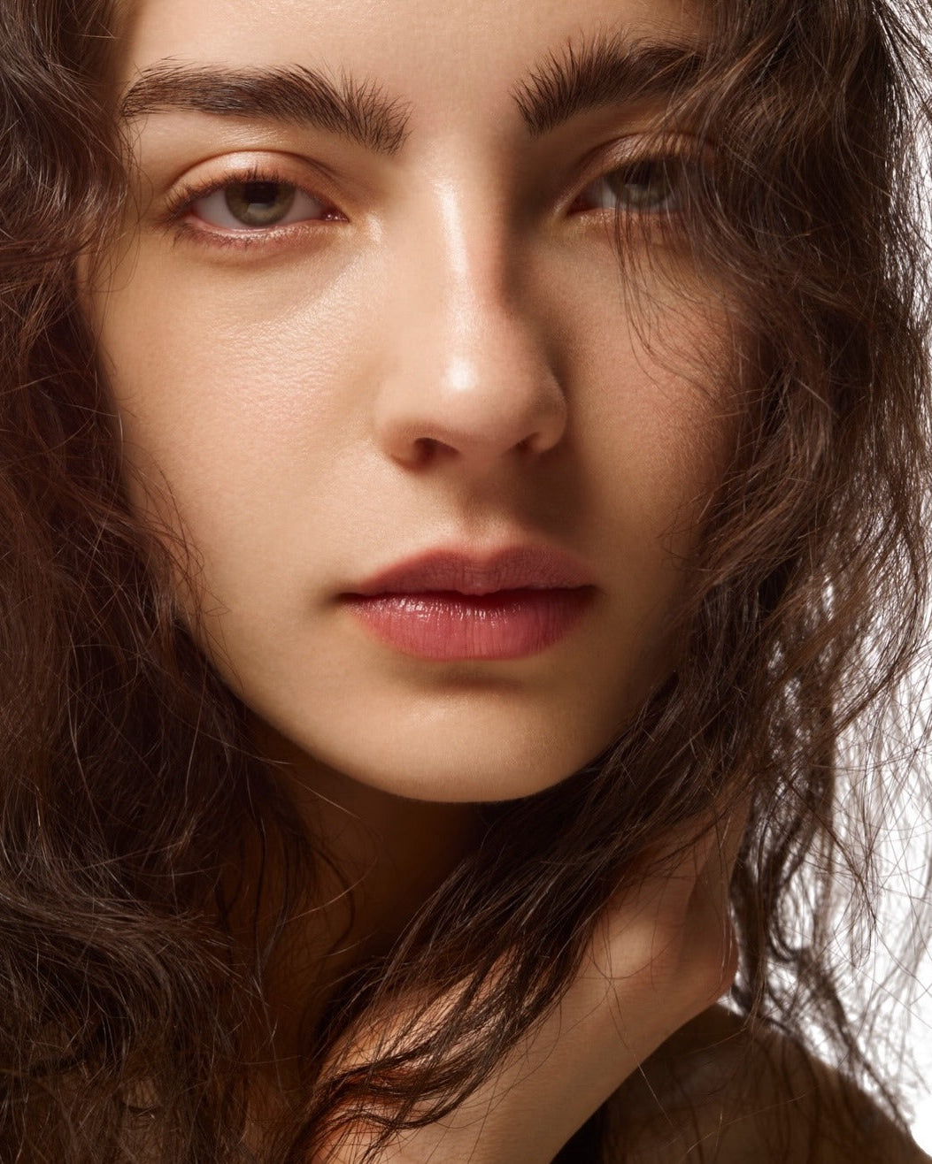 Close-up portrait of a woman with dark brown wavy hair, neutral expression, and soft makeup against a light gray background.